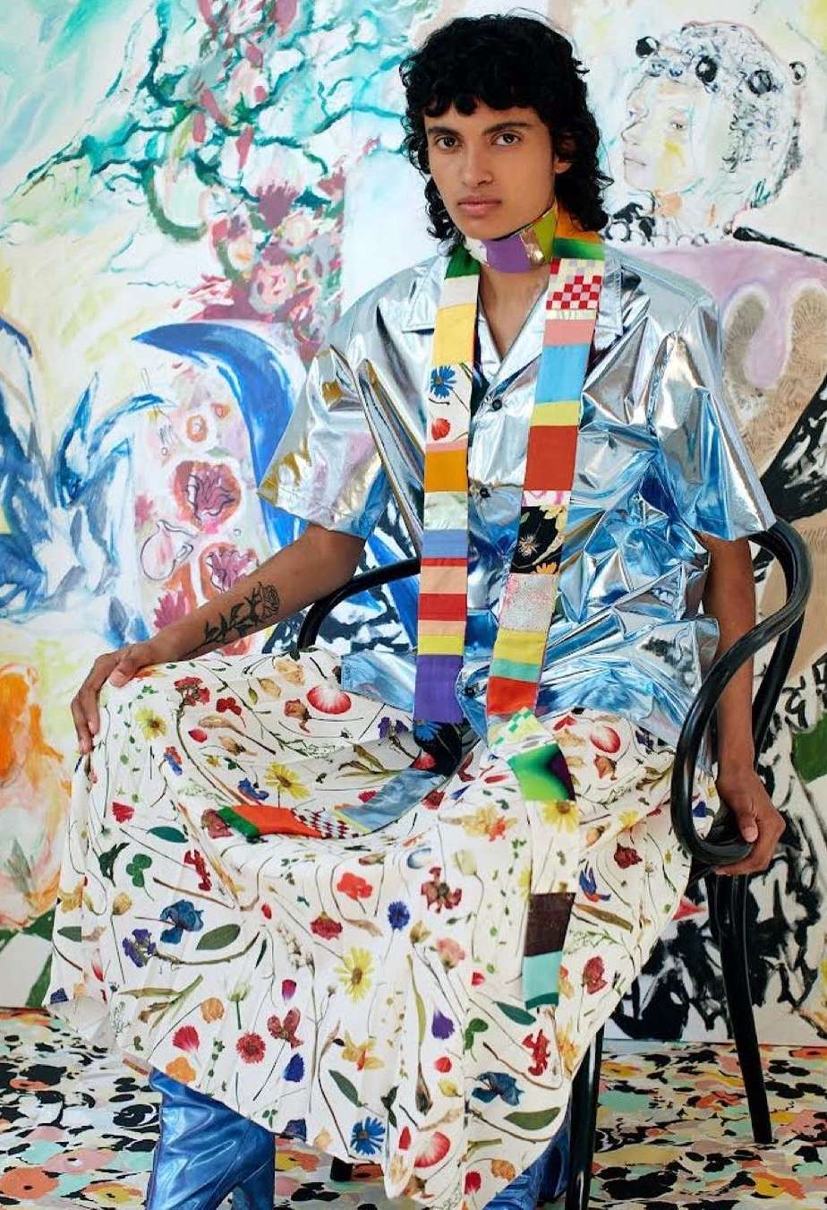 Person sits in avant-garde fashion: shiny shirt, floral skirt, patterned scarf, blue boots. Abstract art backdrop adds a creative flair.