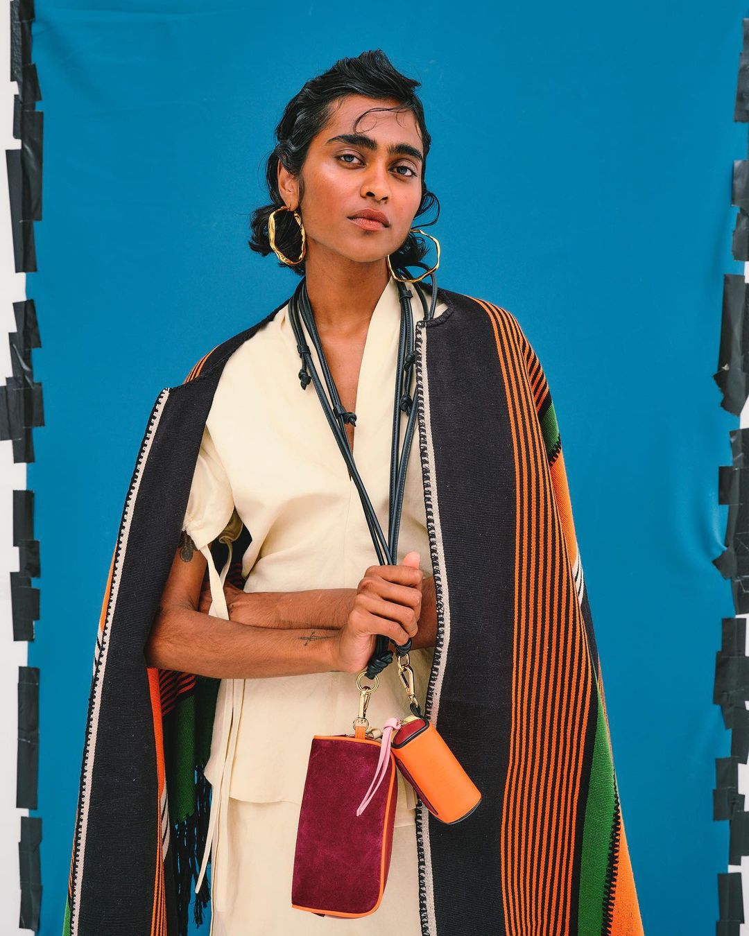 A woman in light-colored clothing stands against a blue backdrop, wearing gold hoop earrings, with a fringed blanket over her shoulders.