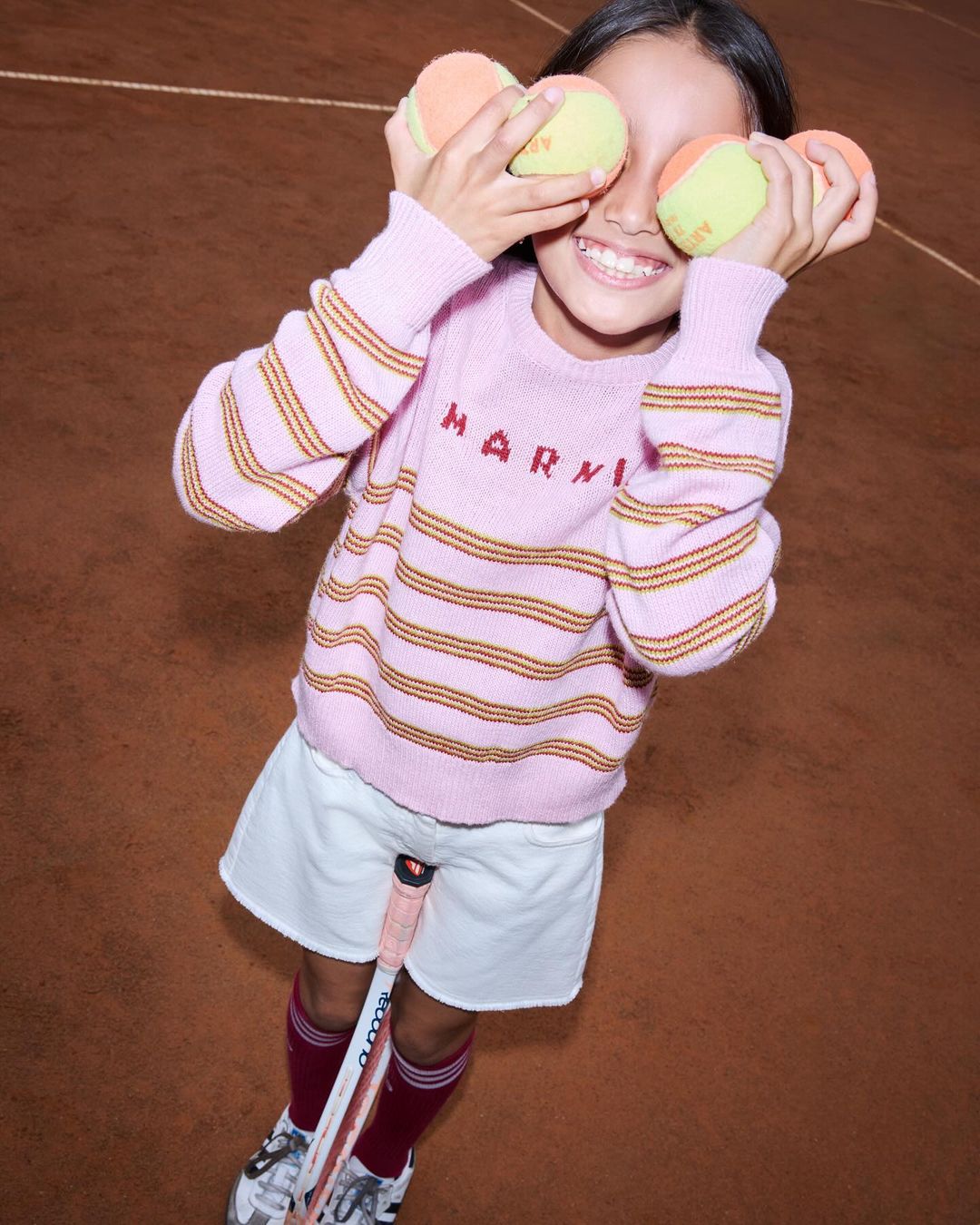 A smiling girl in a pink striped sweater covers her eyes with tennis balls on a brown tennis court, holding a tennis racket.