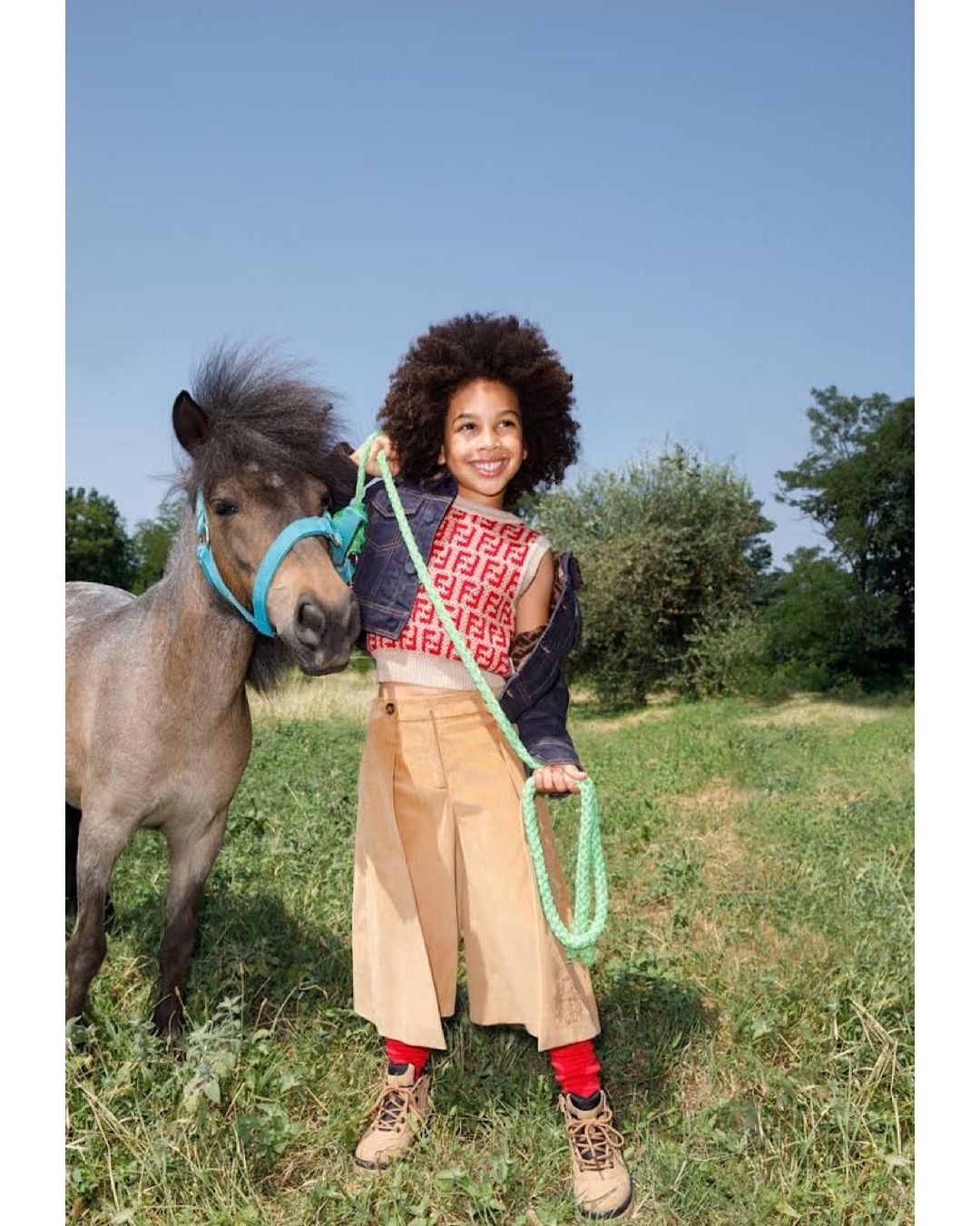A child smiles while holding the reins of a small pony in a grassy field under a clear blue sky, creating a cheerful, outdoor scene.