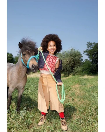 A child smiles while holding the reins of a small pony in a grassy field under a clear blue sky, creating a cheerful, outdoor scene.