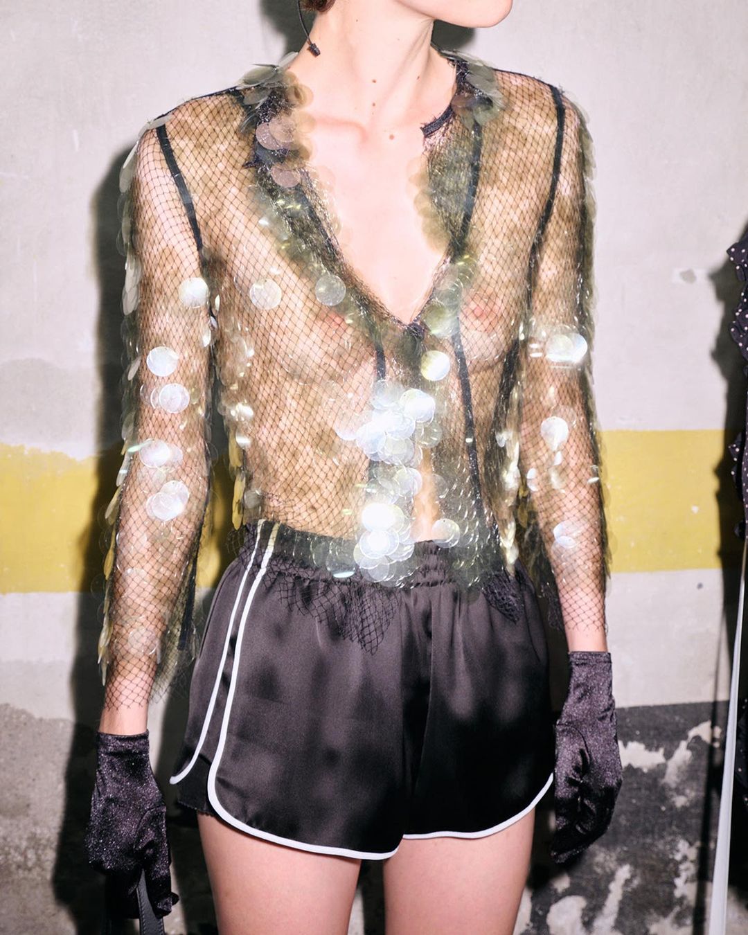 Fashionable adult female wearing a sheer, sequined top and black shorts in a studio with neutral background, emphasizing style and design.
