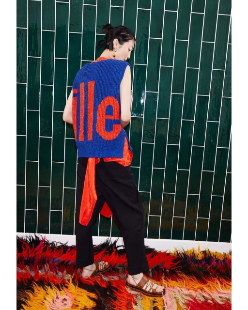 Back view of a woman with a top bun, wearing a blue sweater vest with orange letters, black pants, sandals, and standing against a green tile wall.