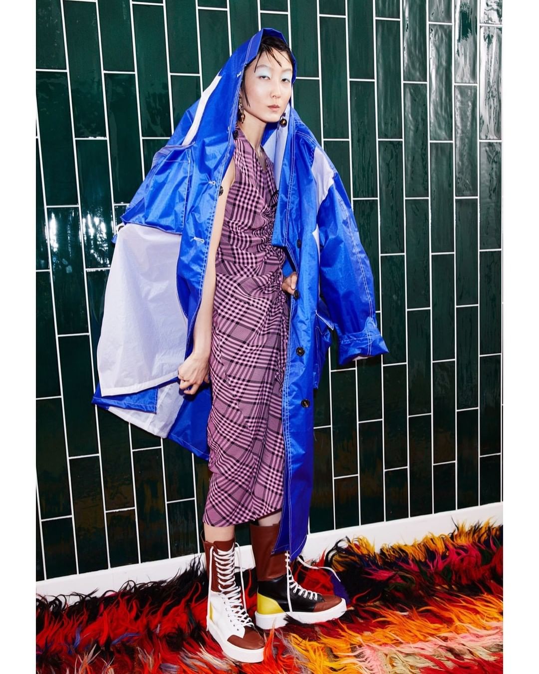 Fashion shot of an Asian woman wearing a dress and a blue hooded jacket, standing on a colorful rug against a green tile wall.