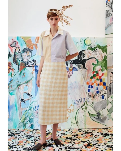 Young woman poses in a mismatched pastel short-sleeve shirt and plaid skirt, standing in front of a colorful abstract painting.