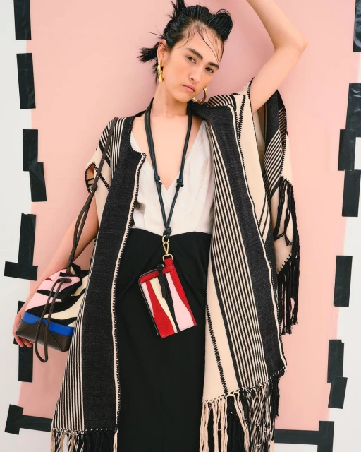 Fashionable woman strikes a pose in a studio setting, wearing a beige and black striped cape, black dress, jewelry, and accessorized with a patterned bag.