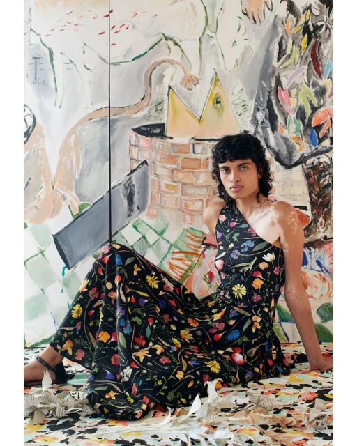 Woman in floral one-shoulder dress sitting in front of a colorful, abstract painting with neutral and brightly painted colors.