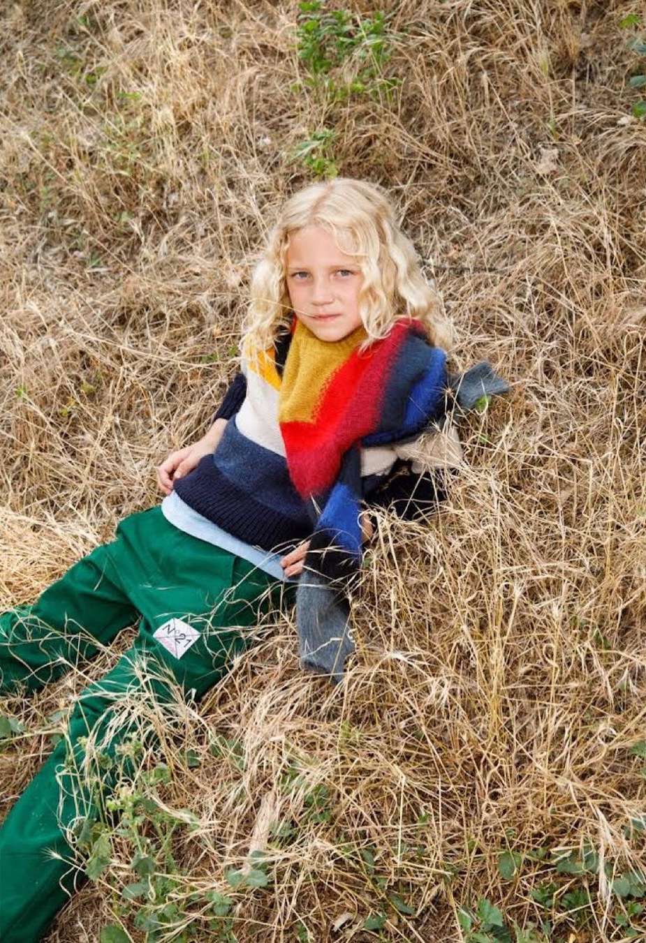 Young child with blond hair lounges outdoors in green pants and a colorful scarf, surrounded by tall, dry grass in a relaxed pose.