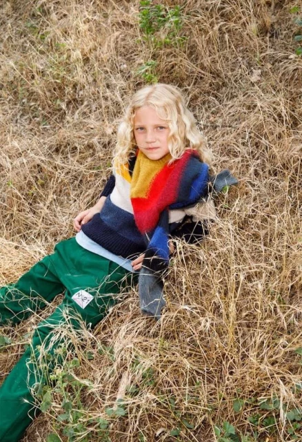 Young child with blond hair lounges outdoors in green pants and a colorful scarf, surrounded by tall, dry grass in a relaxed pose.