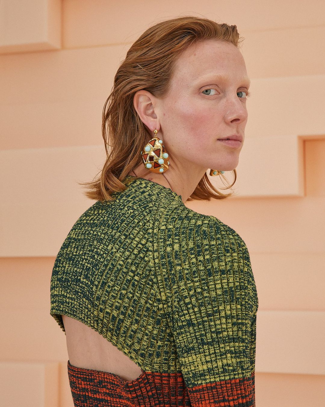 A fashion portrait of a woman with red hair wearing statement earrings and a green and orange textured dress in a studio setting.