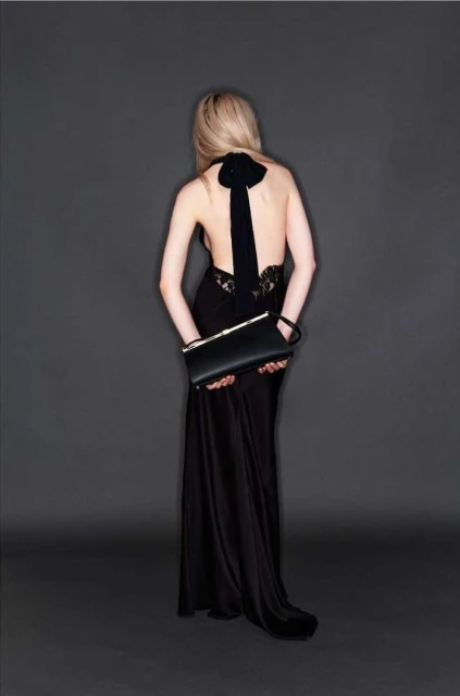 A woman with blond hair, wearing a black gown with a bow, holds a black clutch in a studio setting, photographed from behind.