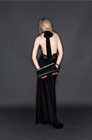 A woman with blond hair, wearing a black gown with a bow, holds a black clutch in a studio setting, photographed from behind.