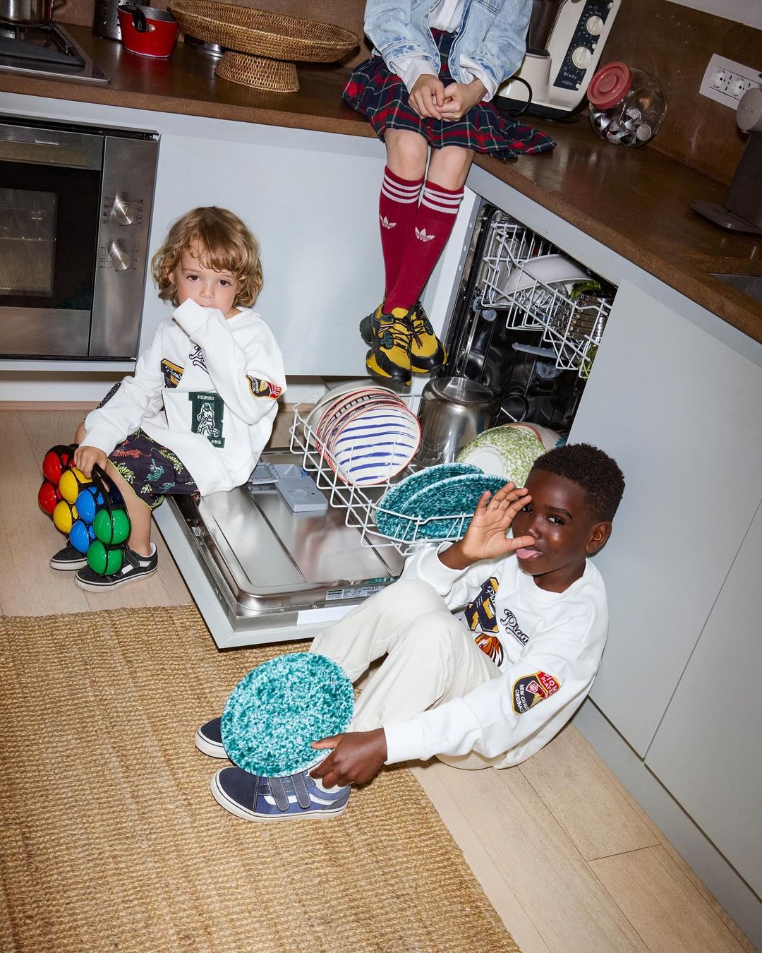 Three young children in a kitchen setting, one seated on a counter, the others on the floor. Casual wear, indoor setting.