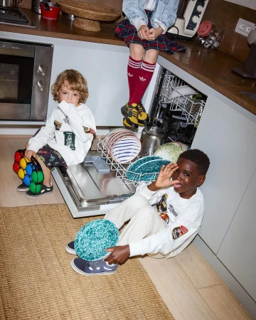 Three young children in a kitchen setting, one seated on a counter, the others on the floor. Casual wear, indoor setting.