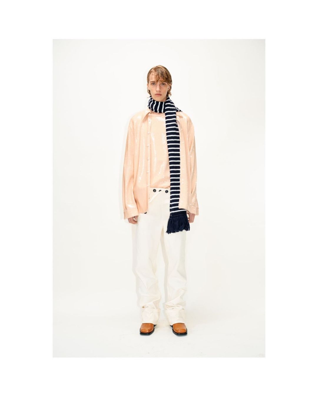 Male fashion model in light pink shirt and white pants, accented by a striped scarf, posing in a bright, neutral photo studio setting.