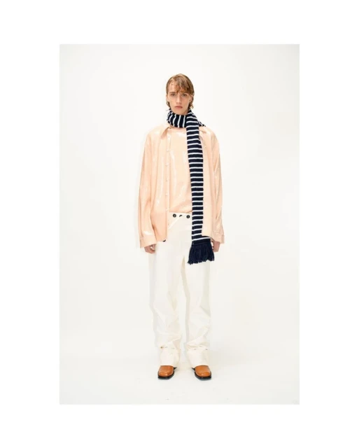 Male fashion model in light pink shirt and white pants, accented by a striped scarf, posing in a bright, neutral photo studio setting.