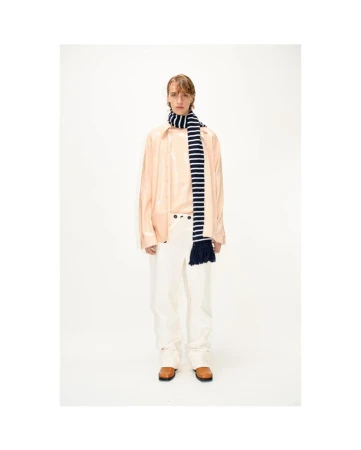 Male fashion model in light pink shirt and white pants, accented by a striped scarf, posing in a bright, neutral photo studio setting.