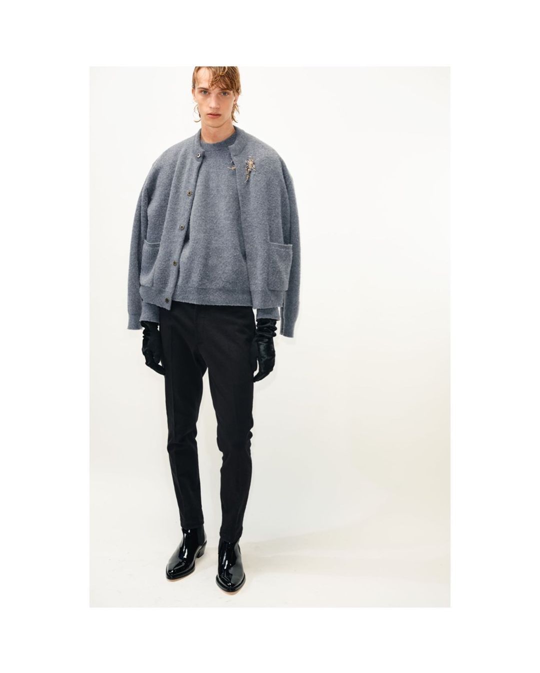 A slender adult male poses in a gray cardigan sweater, black pants, and patent leather boots against a white background in a studio shot.