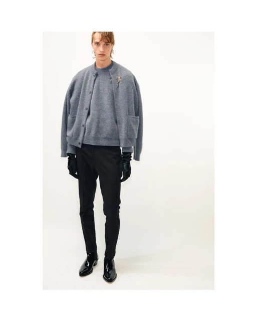A slender adult male poses in a gray cardigan sweater, black pants, and patent leather boots against a white background in a studio shot.