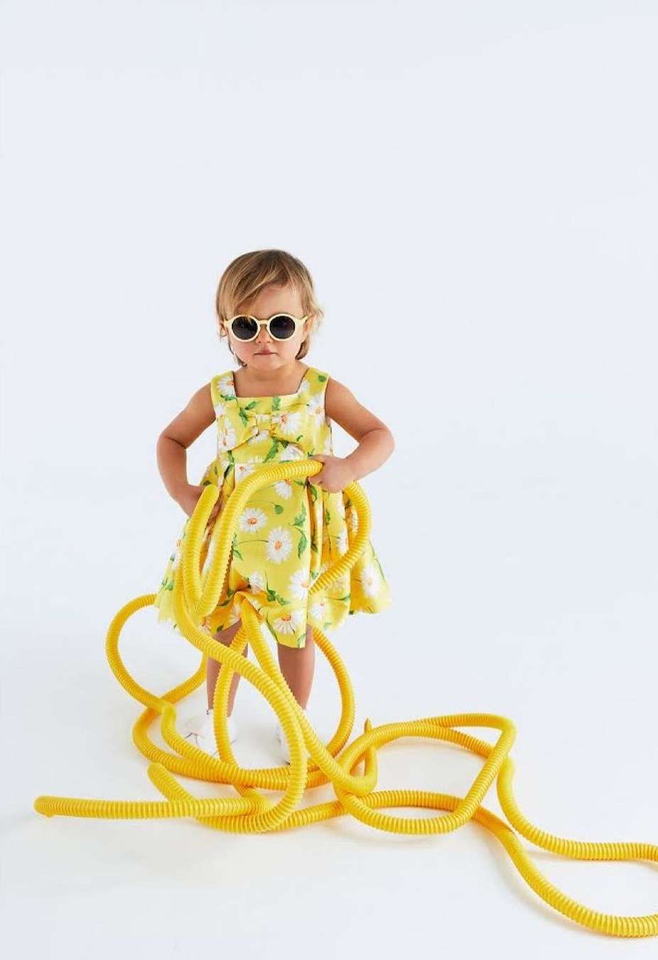 Toddler girl in floral dress and sunglasses poses with a yellow corrugated tube in a bright, high-key studio environment.