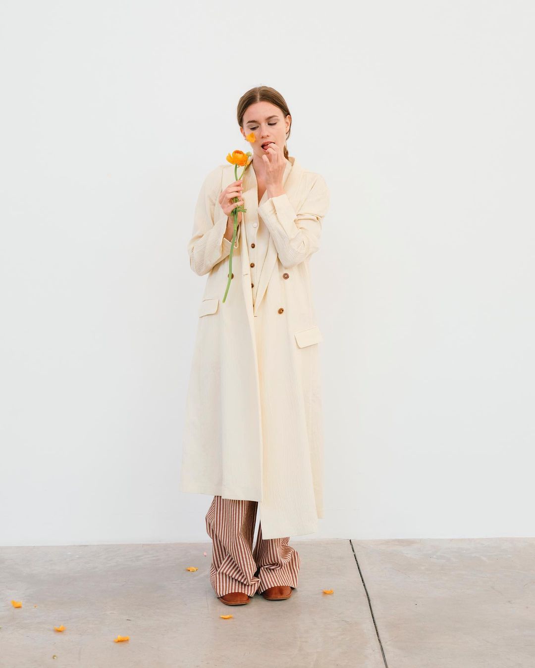 A young woman in a long coat poses against a white wall, holding an orange flower to her face in a minimalist fashion shot.