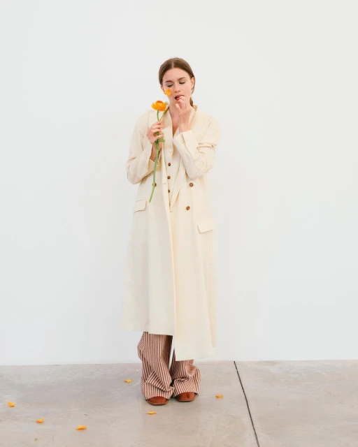 A young woman in a long coat poses against a white wall, holding an orange flower to her face in a minimalist fashion shot.