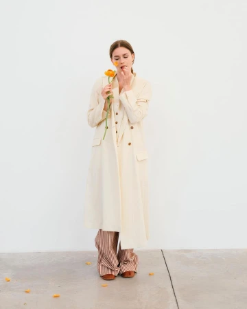 A young woman in a long coat poses against a white wall, holding an orange flower to her face in a minimalist fashion shot.