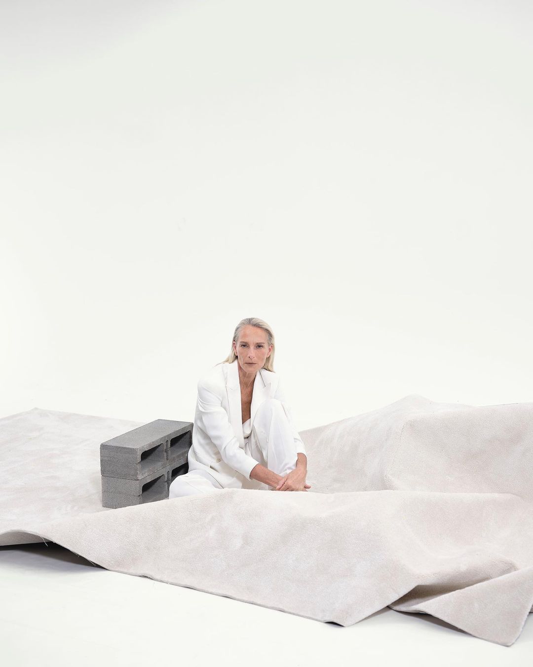 Woman in white suit sitting on a beige carpet next to gray cinder blocks in a white studio with soft, neutral lighting.