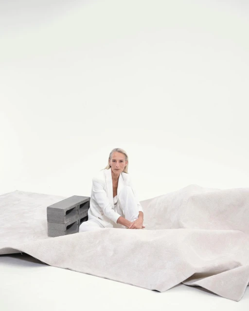 Woman in white suit sitting on a beige carpet next to gray cinder blocks in a white studio with soft, neutral lighting.