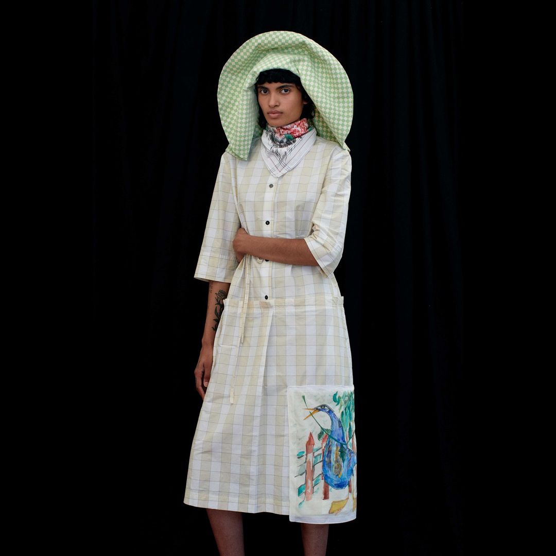 Woman posing in a studio wearing a plaid dress and large hat with a black background, creating a high-fashion and editorial aesthetic.