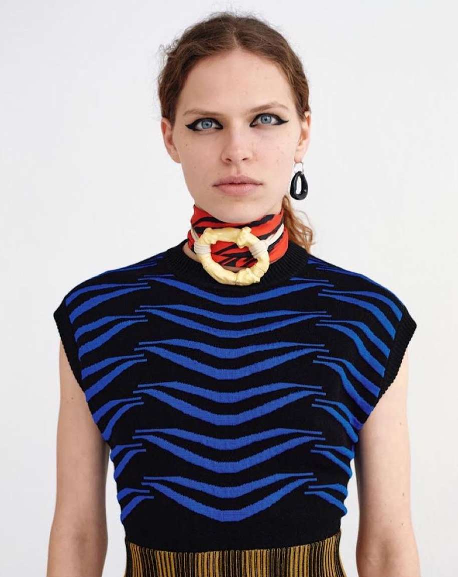 Stylish portrait of a young woman with dark eyeliner, wearing a black and blue wave-patterned top and an eye-catching scarf necklace.