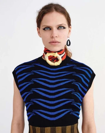 Stylish portrait of a young woman with dark eyeliner, wearing a black and blue wave-patterned top and an eye-catching scarf necklace.