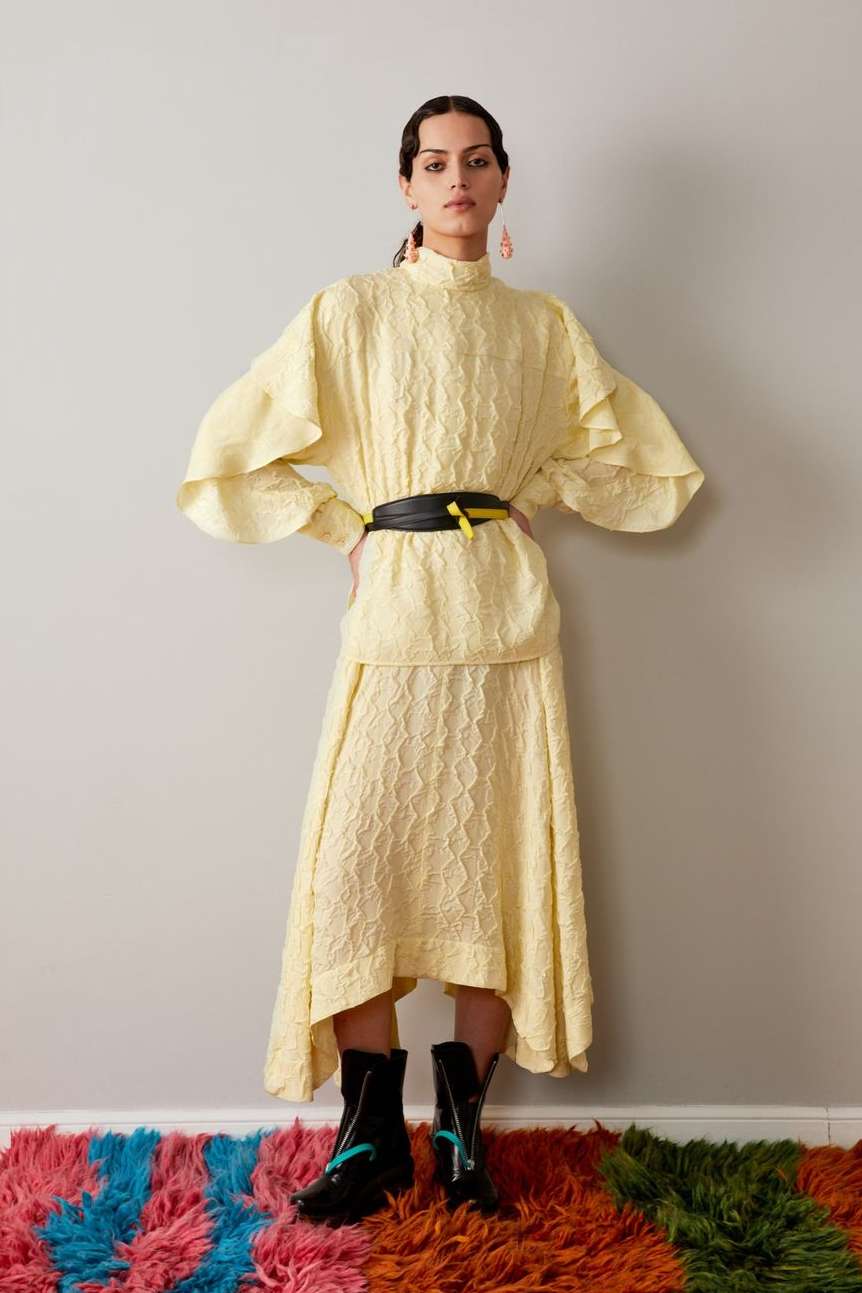 A young adult female model poses in a pale yellow, textured, two-piece dress with a black belt, accessorized with black boots and pink earrings.