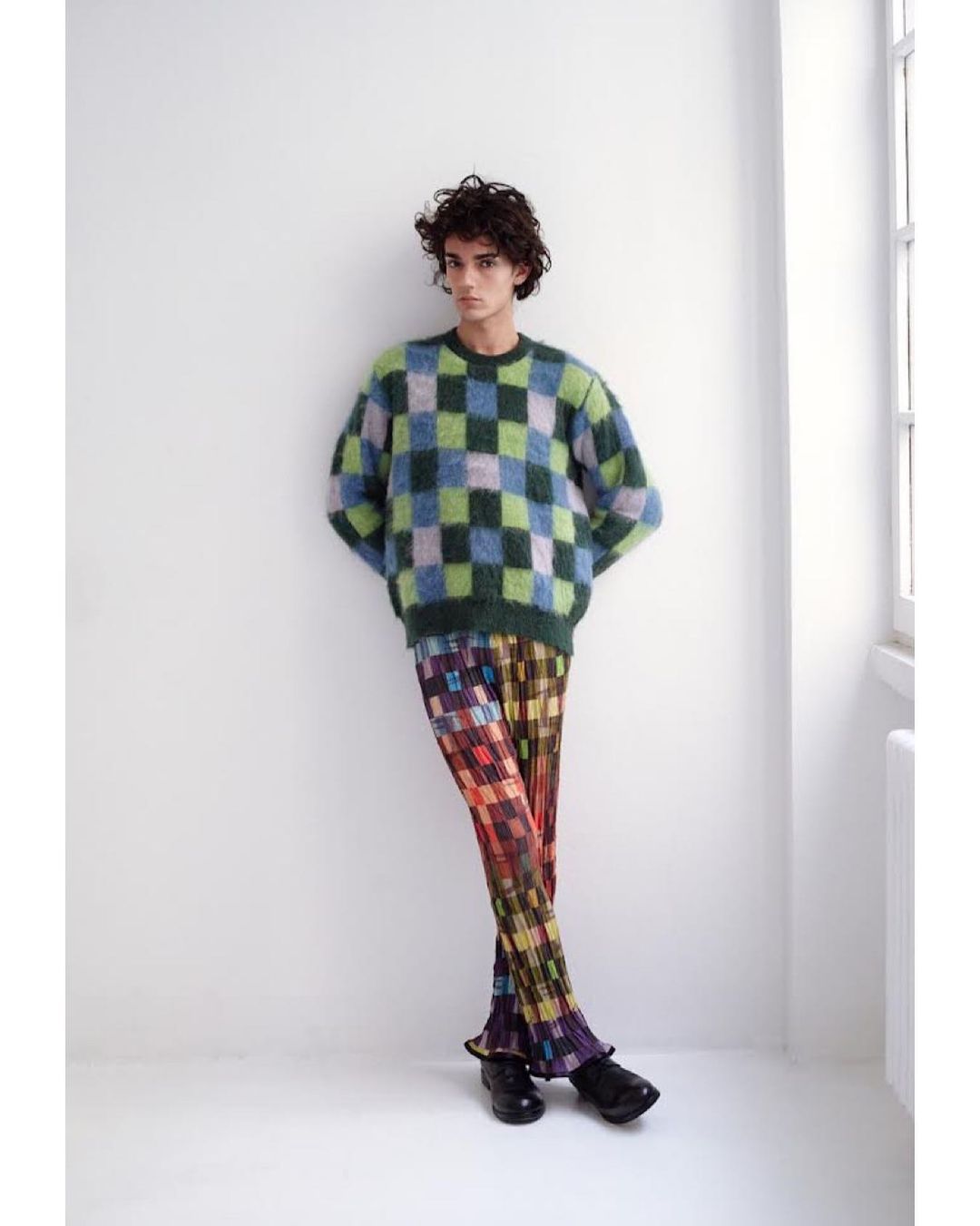 Full-length shot of a curly-haired male model wearing a blue and green checkerboard sweater and multi-colored checkered trousers in studio.