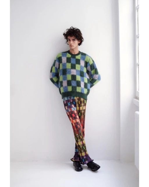 Full-length shot of a curly-haired male model wearing a blue and green checkerboard sweater and multi-colored checkered trousers in studio.