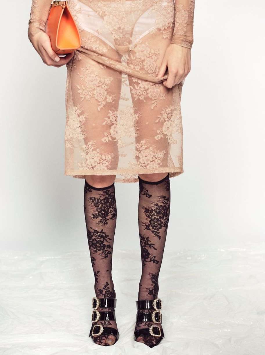 Fashionable woman wearing a lace dress, floral patterned stockings, and buckled heels, holding an orange purse against a white backdrop.