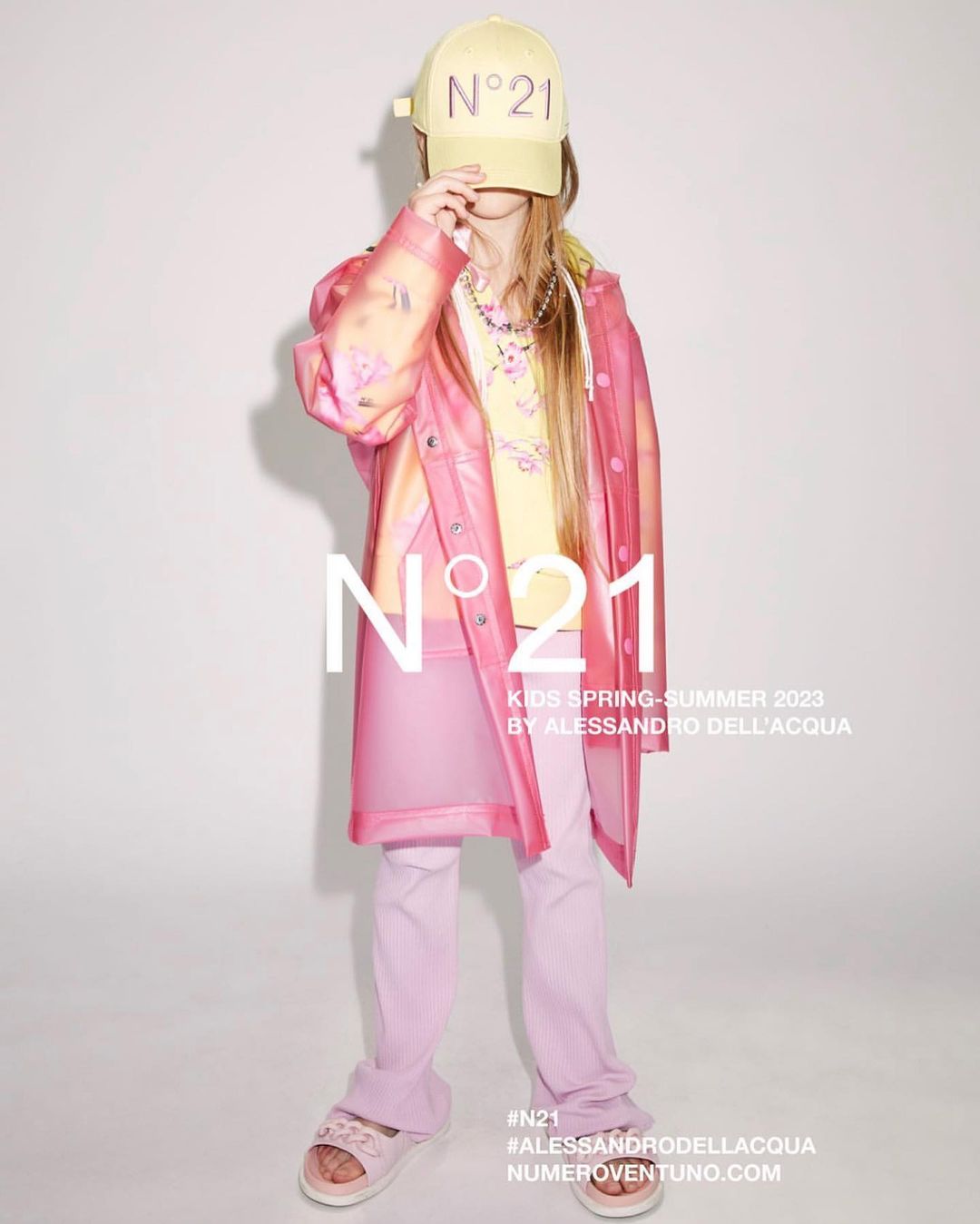 Child fashion shoot for N°21 features a young girl in a yellow baseball cap, printed top, pink translucent jacket and trousers against a white backdrop.