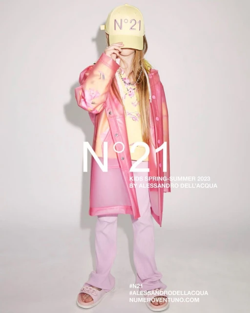Child fashion shoot for N°21 features a young girl in a yellow baseball cap, printed top, pink translucent jacket and trousers against a white backdrop.