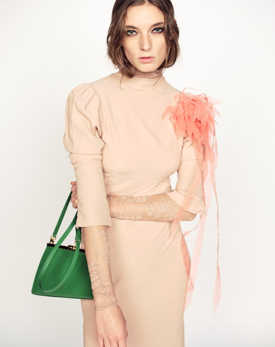 A woman in a light pink dress and lace sleeves poses with a green handbag in a studio setting against a white background.