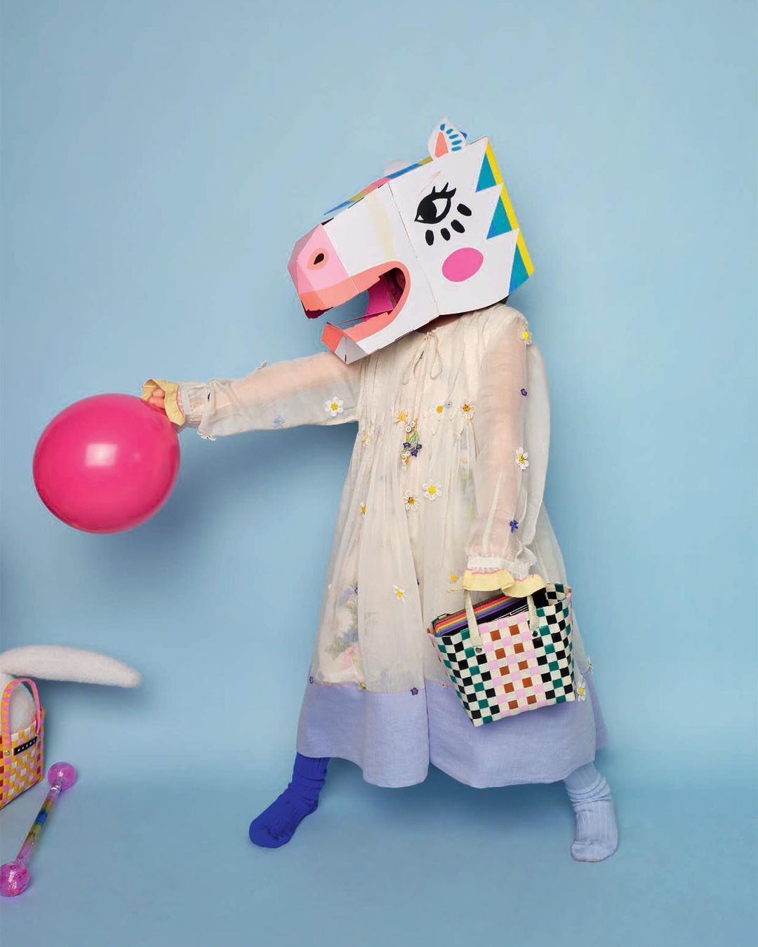 Child in a horse mask stands in front of a blue background, holding a pink balloon and a checker-patterned basket. Studio lighting.