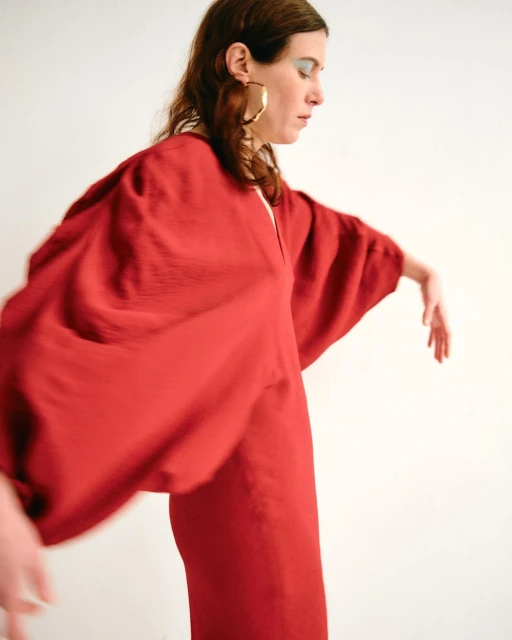Woman in red dress and gold hoop earrings stands in profile, extending arms in studio with white background. Fashion shot, studio lighting.