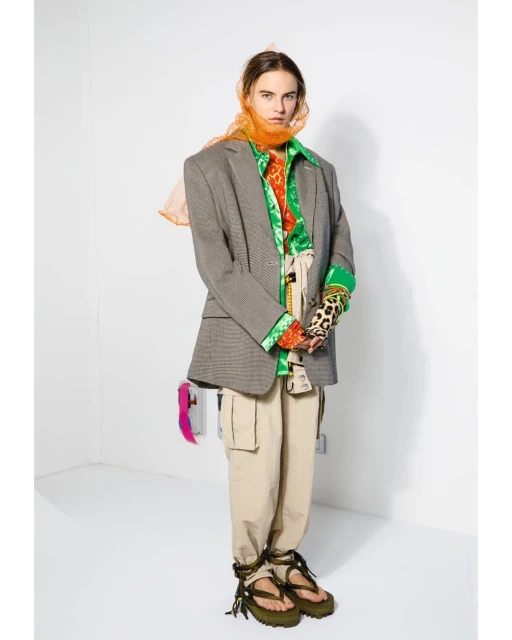 Adult woman wearing a blazer, beige pants, green platform sandals, and an orange scarf posing in a white corner photo studio.