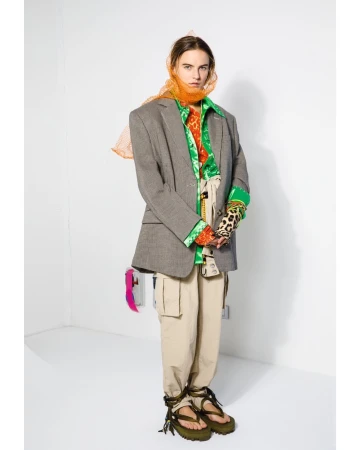 Adult woman wearing a blazer, beige pants, green platform sandals, and an orange scarf posing in a white corner photo studio.