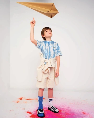 Young boy in tie-dye shirt and shorts points up at a brown paper airplane in a brightly lit photo studio with colorful accents.