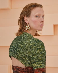 Portrait of a red-haired woman wearing a textured green sweater and statement earrings against a peach studio backdrop.