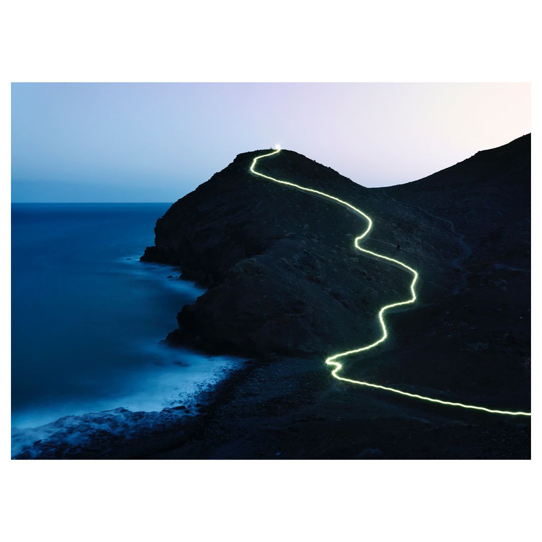 Coastal mountain path illuminated at dusk, light tracing a route up the peak near the ocean under a blue and purple sky.