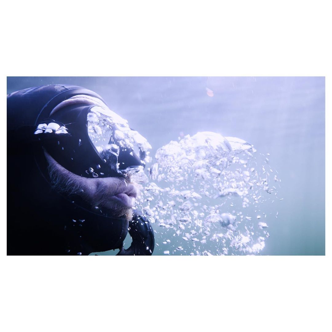 Underwater view of a scuba diver in a dark diving suit exhaling, creating a cluster of bubbles with an aquatic background.