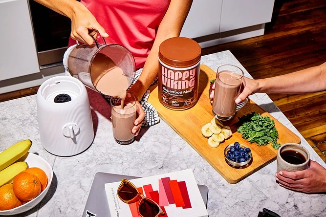 People prepare a chocolate protein smoothie with bananas, blueberries, and kale on a marble countertop with a blender and laptop.