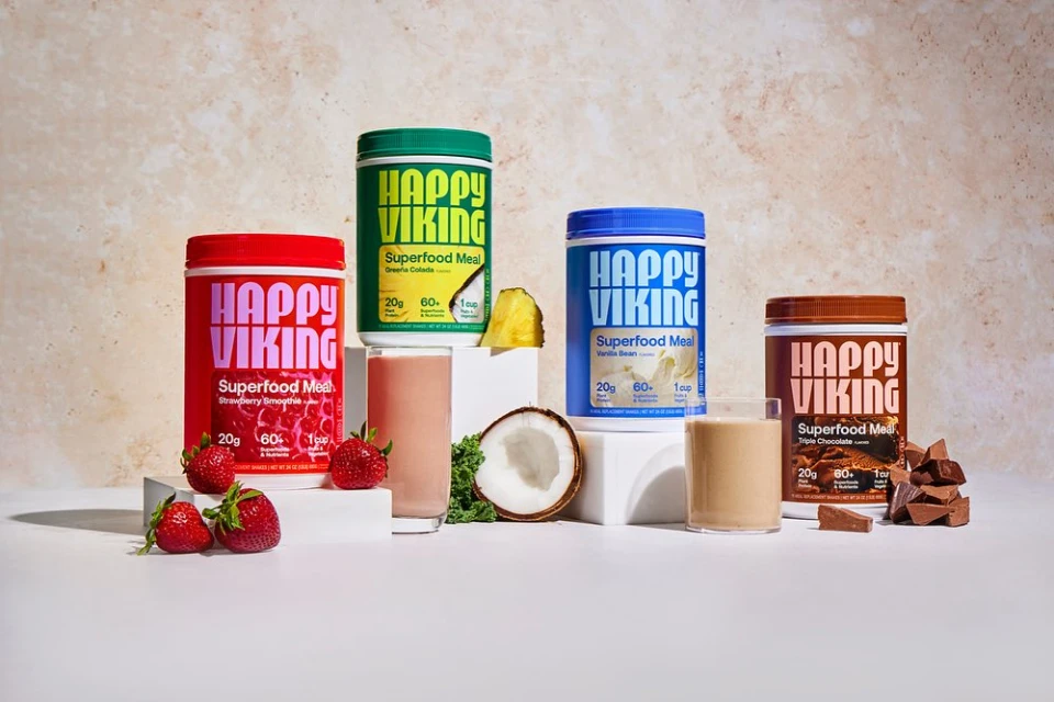 Five containers of Happy Viking Superfood Meal powder sit on a white surface alongside ingredients such as strawberries, pineapple, coconut, and chocolate.