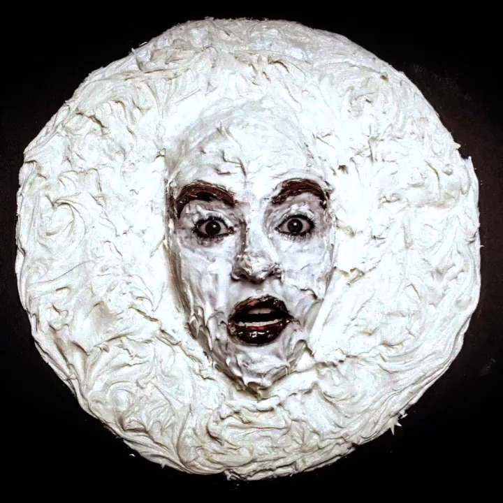 A dramatic portrait features an adult female with white cream covering her face and hair, set against a stark black background.